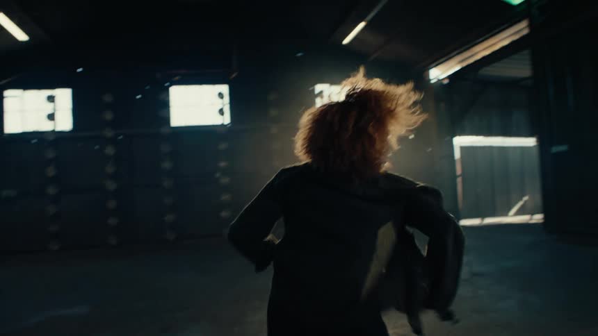 Slow motion of an expressive female dancer with an afro performing a solo contemporary routine in a dark, empty warehouse with dramatic backlighting