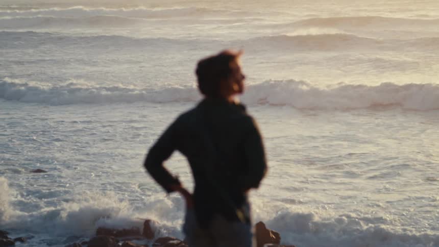 Slow motion view of ocean waves crashing on a rocky shore as an out-of-focus person watches thoughtfully at sunset