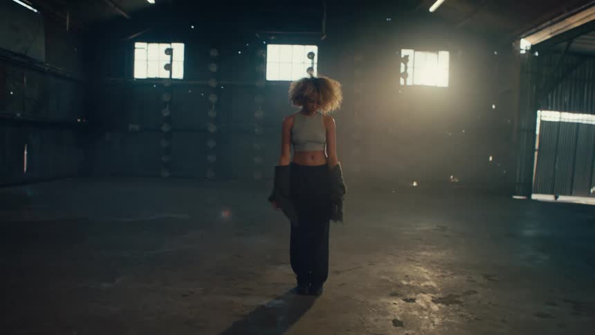 A stylish woman with curly hair dances gracefully in a dusty, sunlit warehouse, moving expressively in slow motion