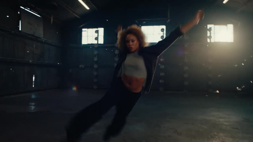 A stylish woman with an afro performs an energetic contemporary dance in a vast, sunlit industrial warehouse