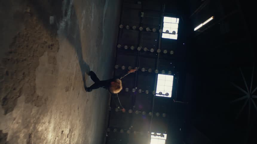 A woman with an afro performs a powerful and expressive dance routine in a vast, sunlit industrial warehouse, captured in dramatic slow motion