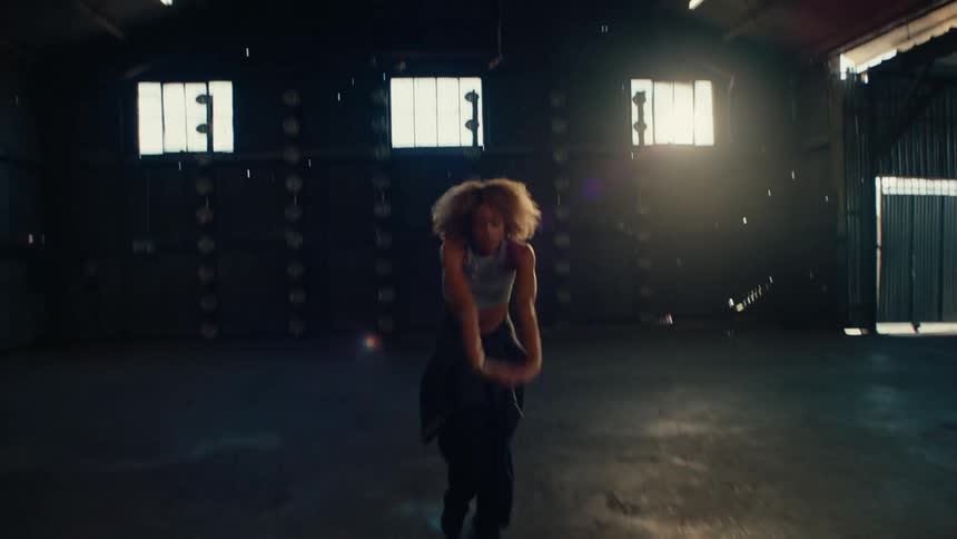 An energetic female dancer with an afro freestyle dances in a large, dark, backlit warehouse space