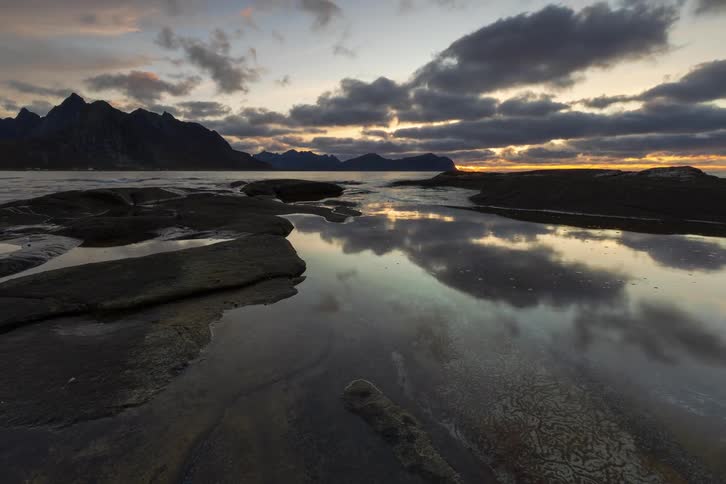 A beautiful timelapse captures the receding tide and a dramatic sunset reflecting off calm waters along a rocky, mountainous coastline