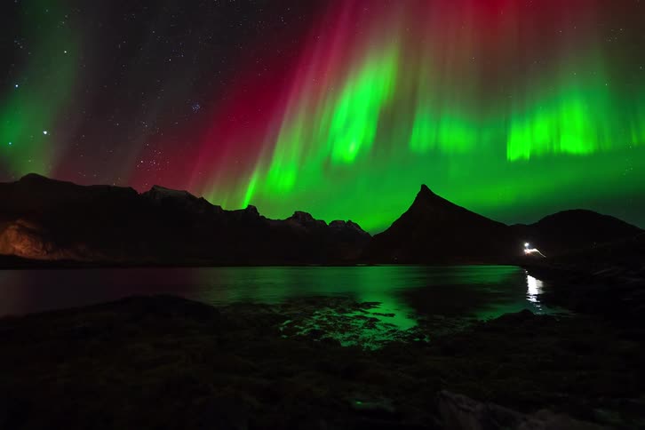 A stunning time-lapse captures the vibrant green and red Aurora Borealis dancing over a majestic mountain range and reflecting in a still lake