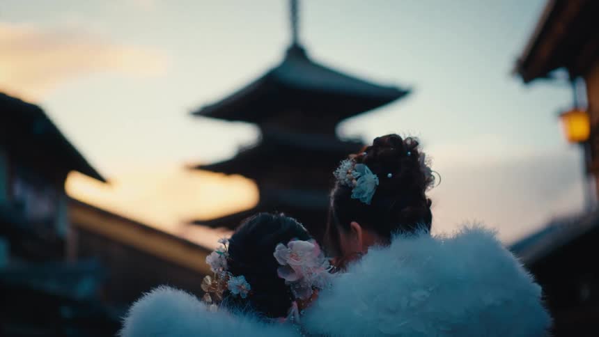 From behind, two women with ornate hairstyles and white fur shawls gaze at a traditional Japanese pagoda silhouetted against the sunset