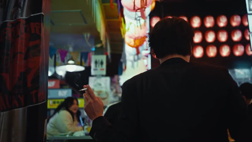 A man seen from behind holds a lit cigarette while standing in a bustling, colorful Asian night market