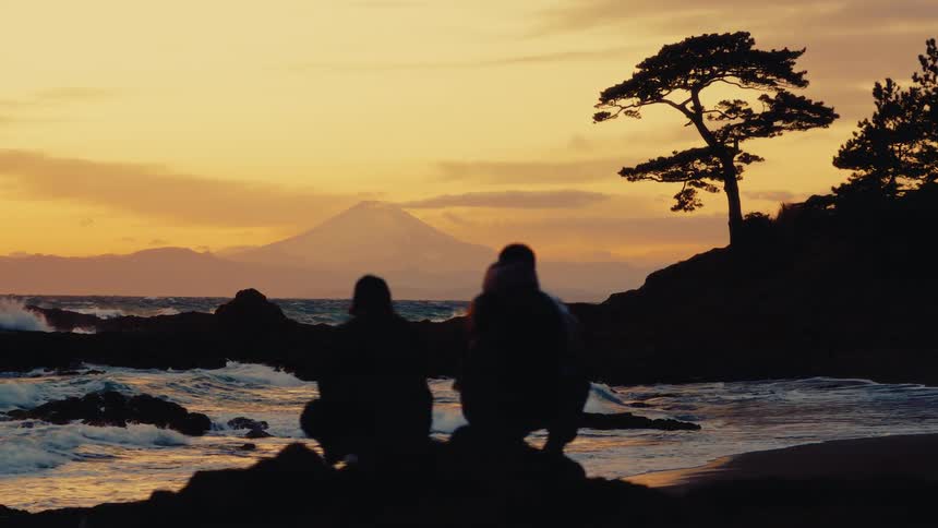A cinemagraph of two silhouetted figures watching the waves crash on a Japanese beach at sunset with Mount Fuji in the distance