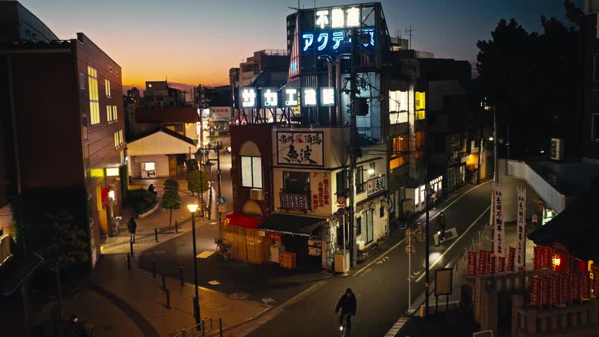 A static high-angle view of a Tokyo street corner with glowing neon signs at dusk as people pass by