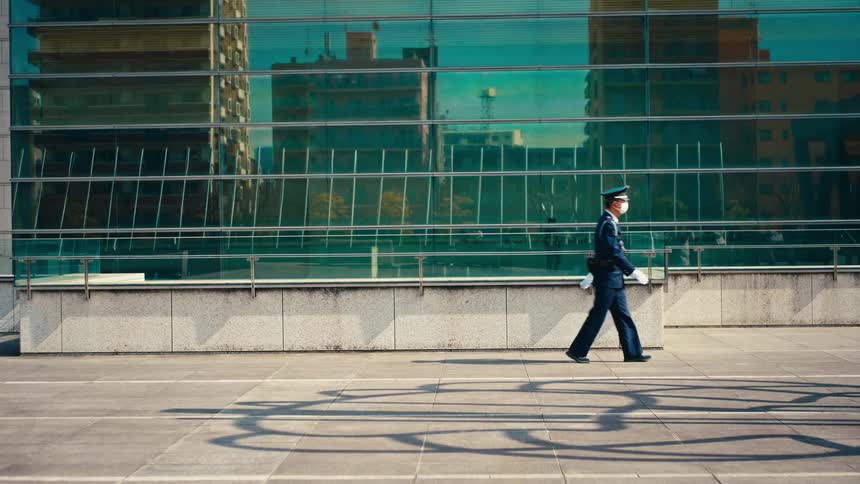 A Japanese police officer wearing a face mask walks past a modern glass building on a sunny city day
