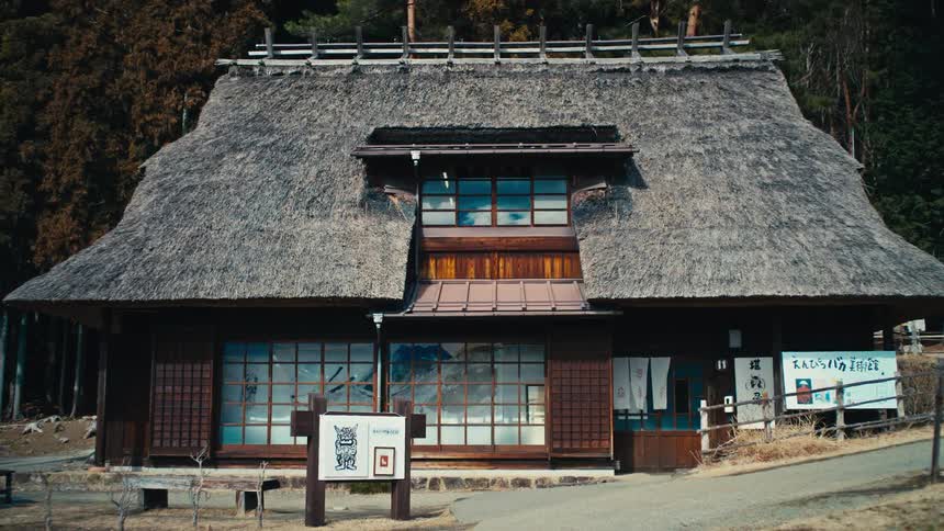 A traditional Japanese farmhouse with a thatched roof sits peacefully as clouds reflect in the windows