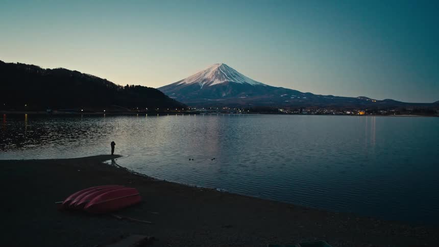 A lone figure stands on the shore of a tranquil lake at dawn, admiring the majestic, snow-capped peak of Mount Fuji in Japan