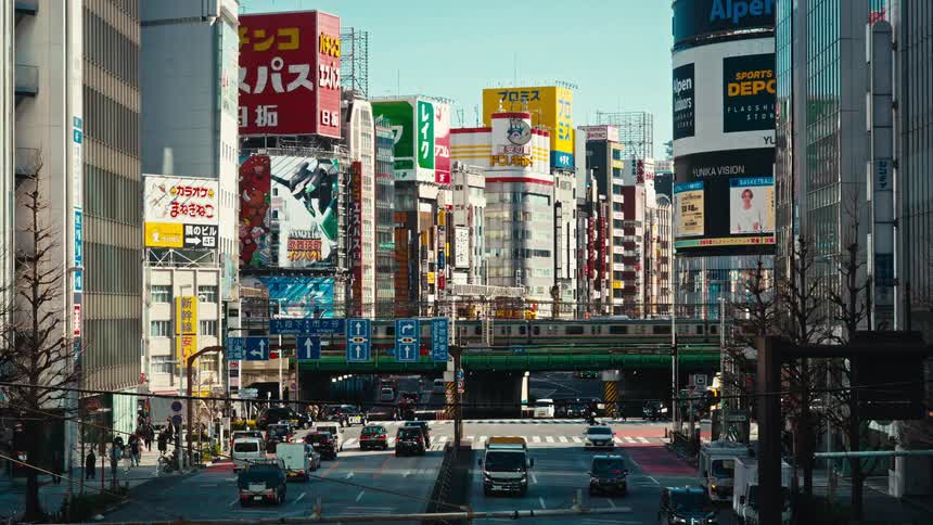 A train passes over a bustling street intersection in Shinjuku, Tokyo, surrounded by towering buildings and vibrant advertisements