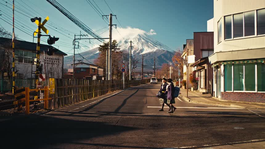 A cyclist and two schoolgirls cross a quiet street in a Japanese town with a stunning view of Mount Fuji looming in the background