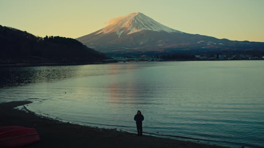 A lone figure stands on the shore of a tranquil lake, gazing at the majestic snow-capped Mount Fuji bathed in the warm light of sunrise