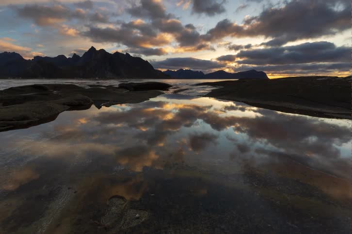 A breathtaking timelapse of a sunset over majestic mountains, with clouds perfectly reflected in the still waters of a rocky shoreline