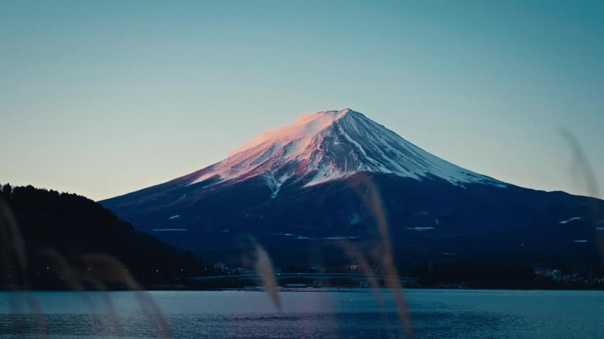 The iconic snow-capped peak of Mount Fuji glows with the pink light of sunrise as seen from across a still lake in Japan