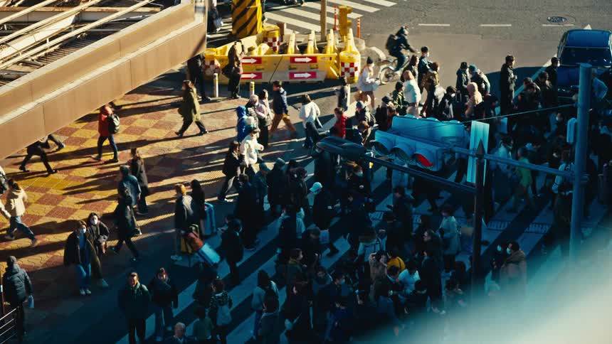 A high-angle slow-motion shot captures a diverse crowd of pedestrians crossing a busy sunlit street in a modern city