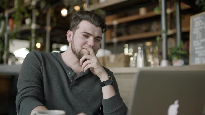 A man sitting at a table looking at a computer screen sits back to laugh
