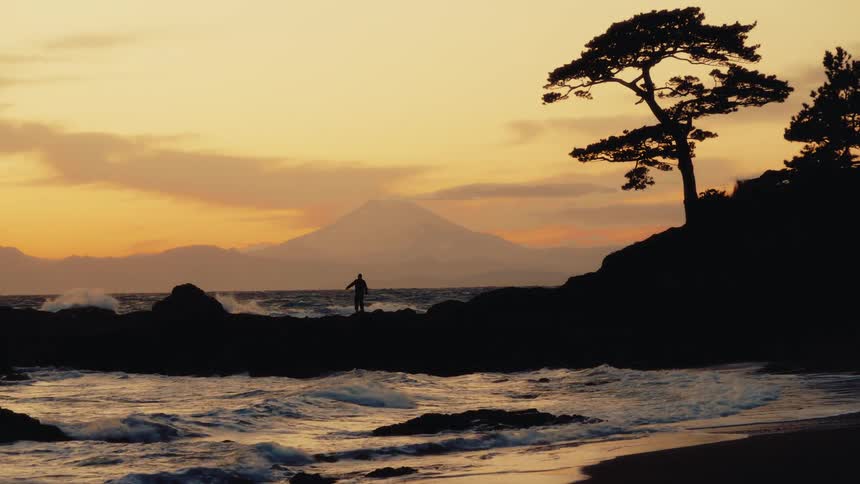 A silhouetted person walks along a rocky shoreline at sunset, with waves rolling in and Mount Fuji visible on the horizon