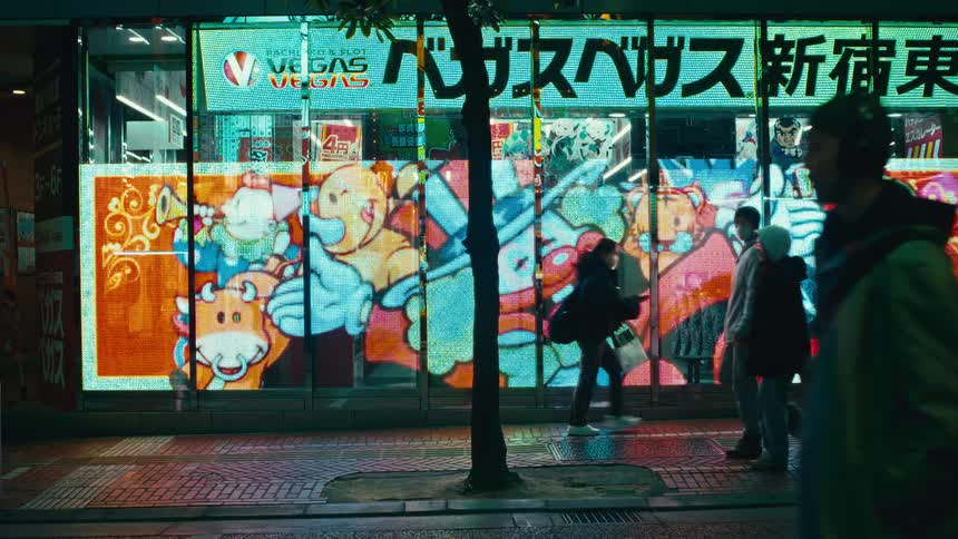 Pedestrians walk past a brightly illuminated storefront with flashing anime-style advertisements on a bustling Tokyo street at night