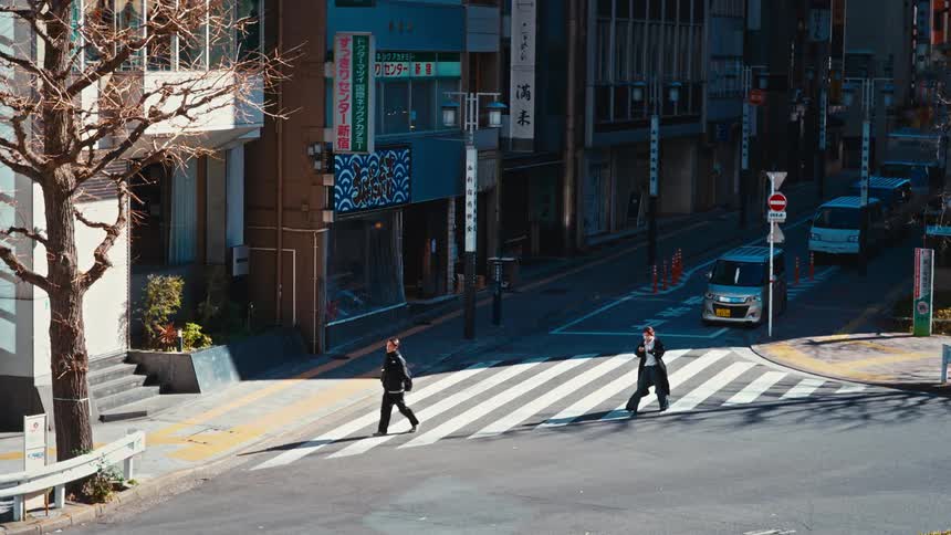 A man and woman walk in opposite directions across a sunny street corner in a Japanese city in cinematic slow motion