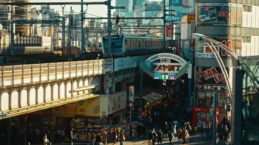 A commuter train passes over the bustling Ameyokocho Market in Ueno, Tokyo, as crowds of pedestrians walk below during the golden hour