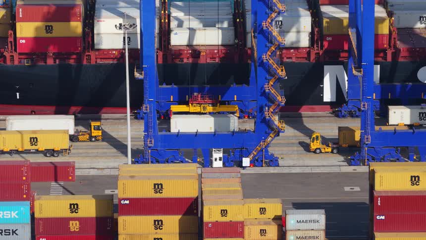 A high-angle view of a large blue gantry crane unloading a container from a cargo ship at a bustling industrial port
