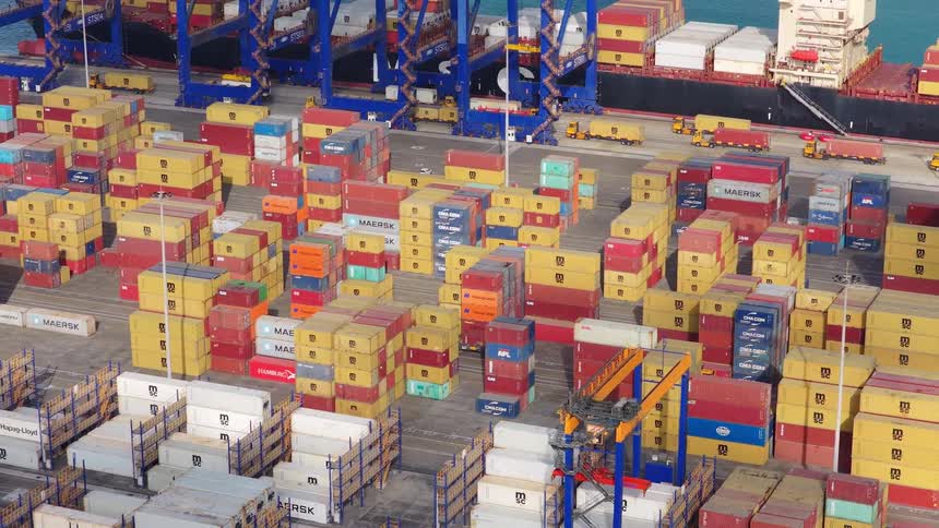 Slowly zooming out aerial shot of a bustling container port, showing stacks of cargo containers, moving trucks, and giant cranes alongside a docked ship