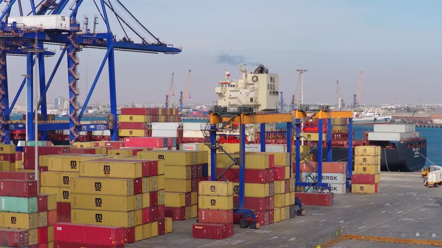 A bustling industrial shipping port shows gantry cranes and forklifts moving colorful cargo containers with a large ship docked in the background