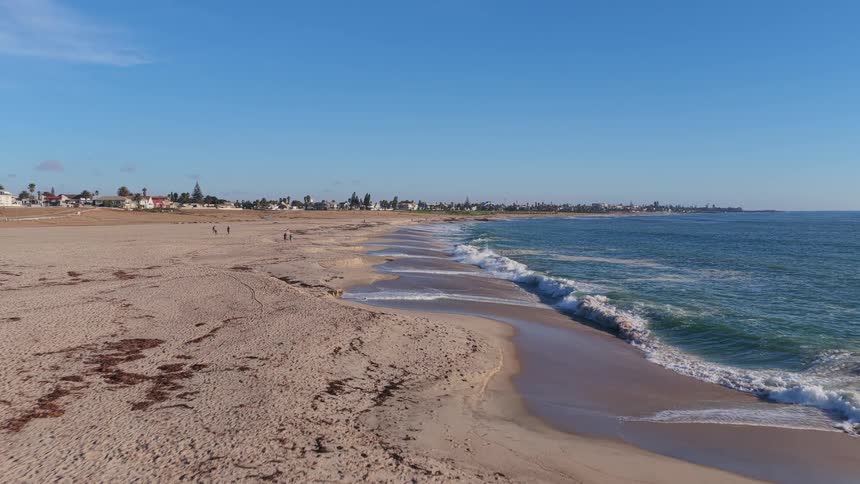 A slow aerial shot moves forward along a vast sandy beach as gentle waves roll onto the shore, with a coastal town visible in the distance
