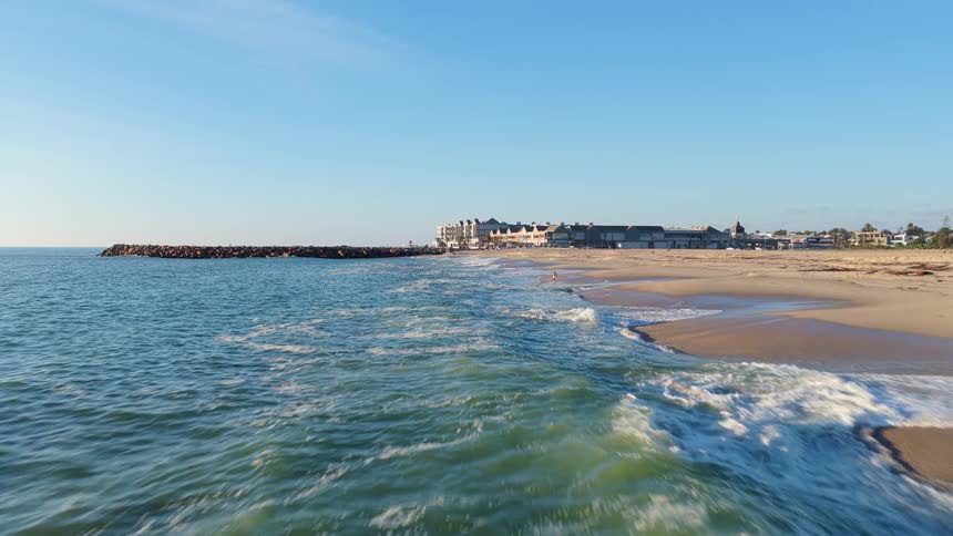 An aerial view of ocean waves gently crashing on a sandy beach with a coastal town in the background on a sunny day