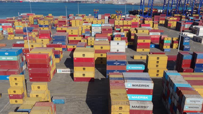 An aerial view slowly advances over a vast and colorful shipping container terminal at a busy industrial port by the sea
