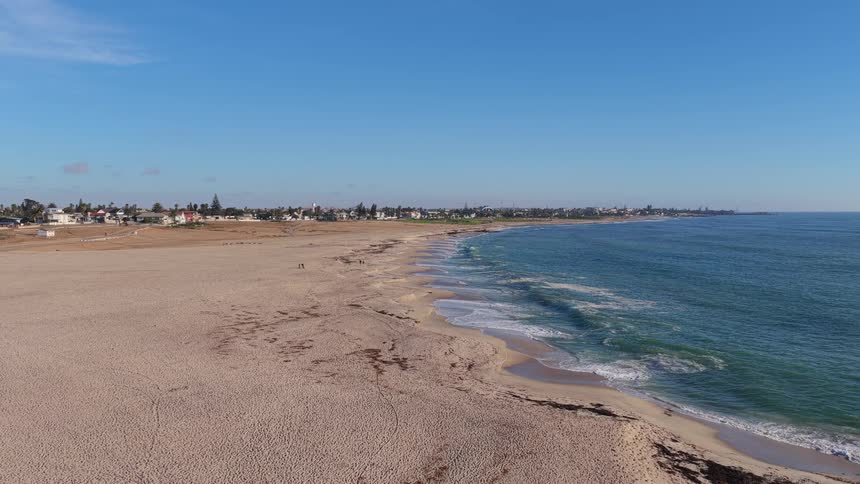 A slow aerial pullback reveals a vast, serene sandy beach where gentle ocean waves meet the shore, with a distant coastal town under a brilliant blue sky