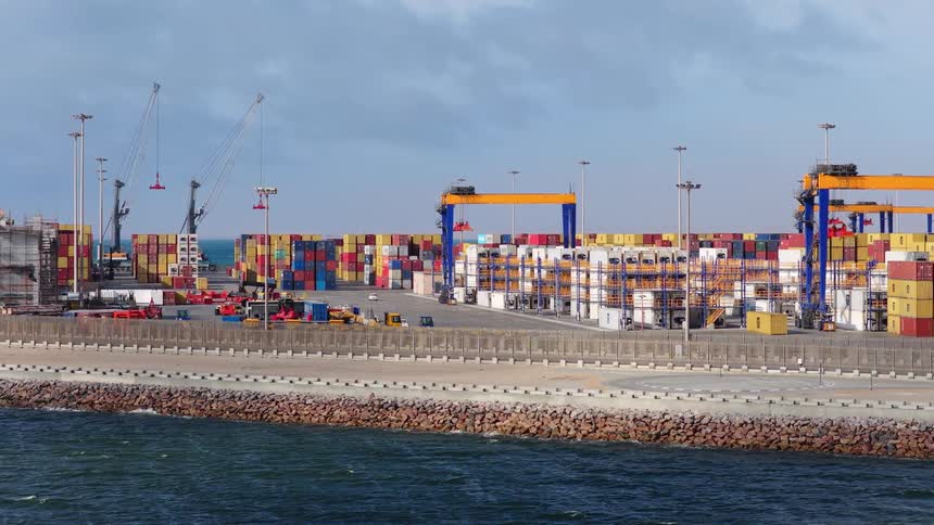 A wide shot of a bustling seaside container port with gantry cranes and trucks moving cargo amongst colorful shipping containers