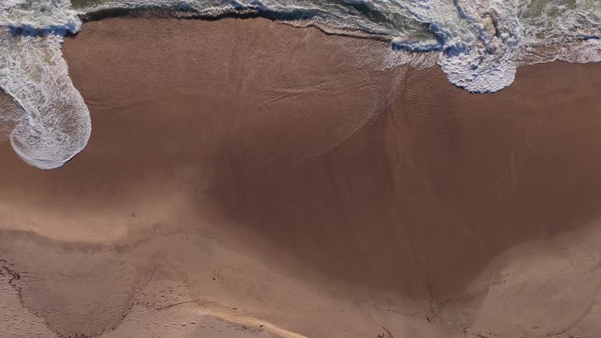 Slow motion aerial footage from directly above shows gentle ocean waves washing onto a sandy beach