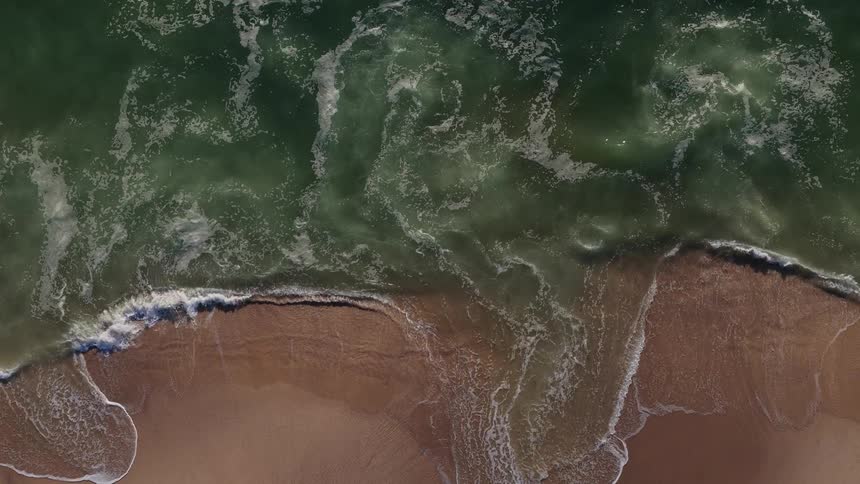 Mesmerizing top-down aerial shot of green ocean waves washing over a sandy beach in slow motion