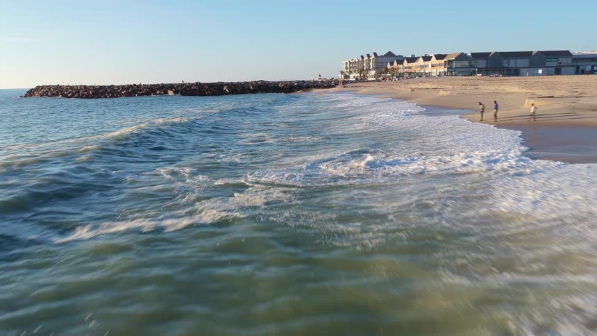 A low altitude aerial shot flies backward over gentle ocean waves breaking on a quiet sandy shore next to a stone jetty