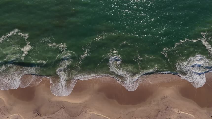 A top-down aerial slow-motion view of turquoise ocean waves gently lapping against a sandy beach