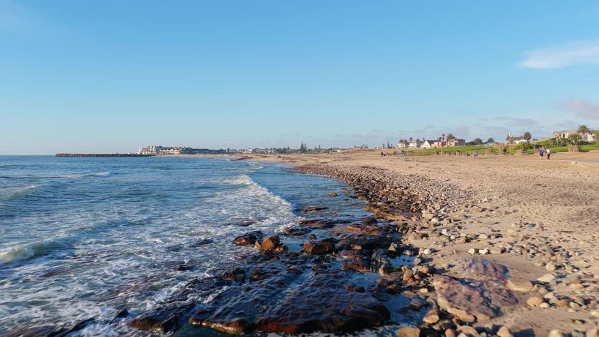 An aerial drone shot slowly flies along a beautiful sandy and rocky coastline as gentle waves wash ashore under a clear blue sky