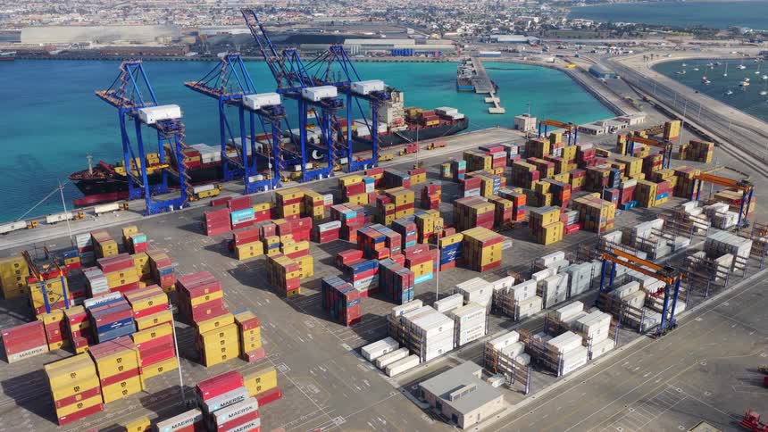 Static aerial view of a bustling shipping port with massive cranes, colorful stacked containers, and a cargo ship docked