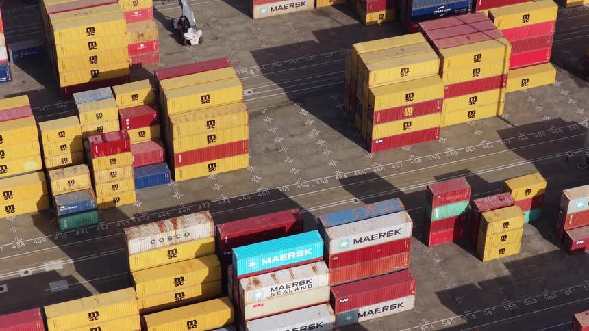 A high-angle static shot captures stacks of colorful shipping containers at a busy industrial port terminal
