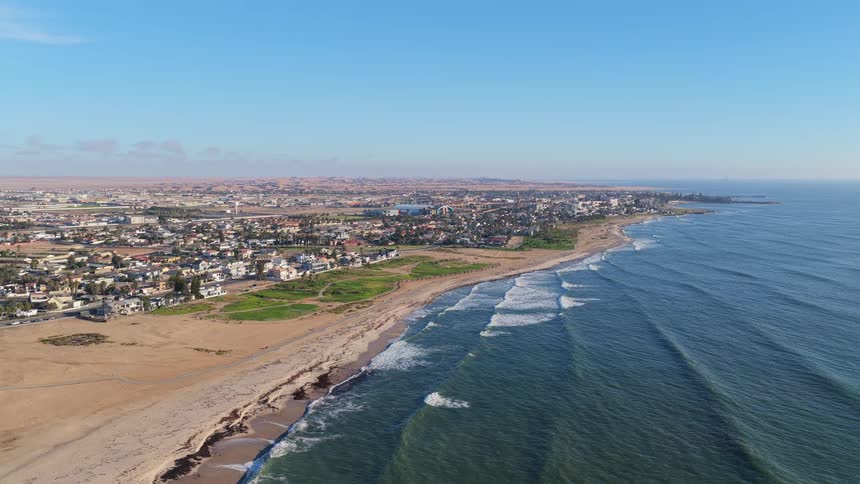 A wide aerial shot shows the Namibian coastal city of Swakopmund, where houses line the shore between the vast desert and the rolling ocean waves
