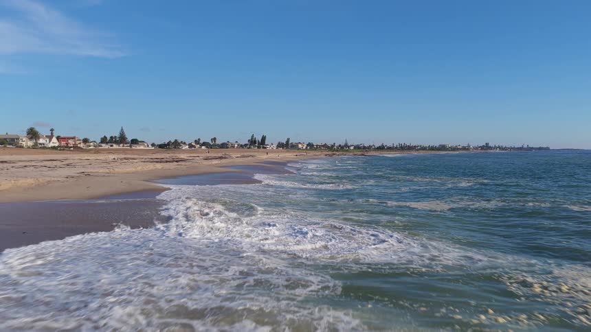 A low-angle, slow-motion shot of gentle ocean waves washing up on a sandy beach with a coastal town in the distance