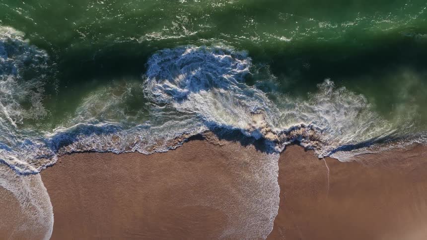 A mesmerizing top-down aerial view of emerald green ocean waves breaking in slow motion onto a sandy shore