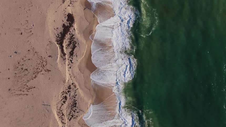 An aerial top-down slow-motion view captures the serene beauty of ocean waves gently lapping against a sandy shore