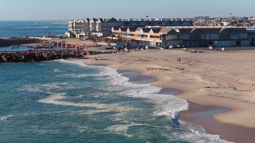 Static aerial view of calm ocean waves washing ashore on a sandy beach next to a small coastal town and marina