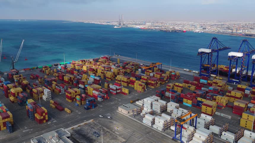 A wide aerial view of a busy shipping container terminal at a port, showcasing global trade and logistics by the sea