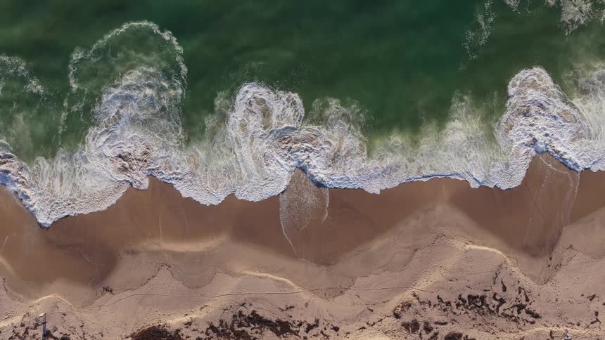 An aerial top-down view of mesmerizing green ocean waves crashing onto a sandy shore in slow motion