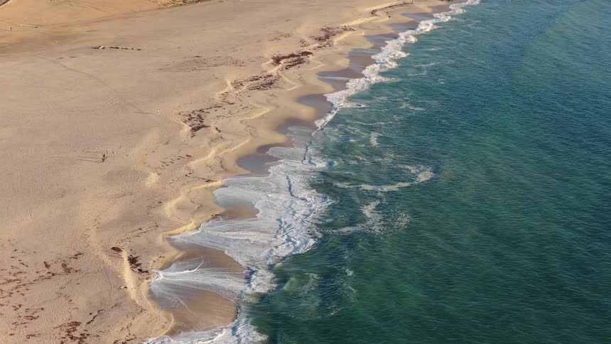An aerial perspective shows tranquil ocean waves breaking on a deserted sandy beach