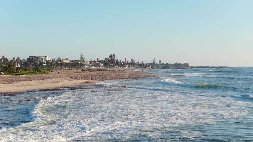 An aerial perspective flies low over the tranquil ocean, moving towards a sandy beach and a coastal town promenade